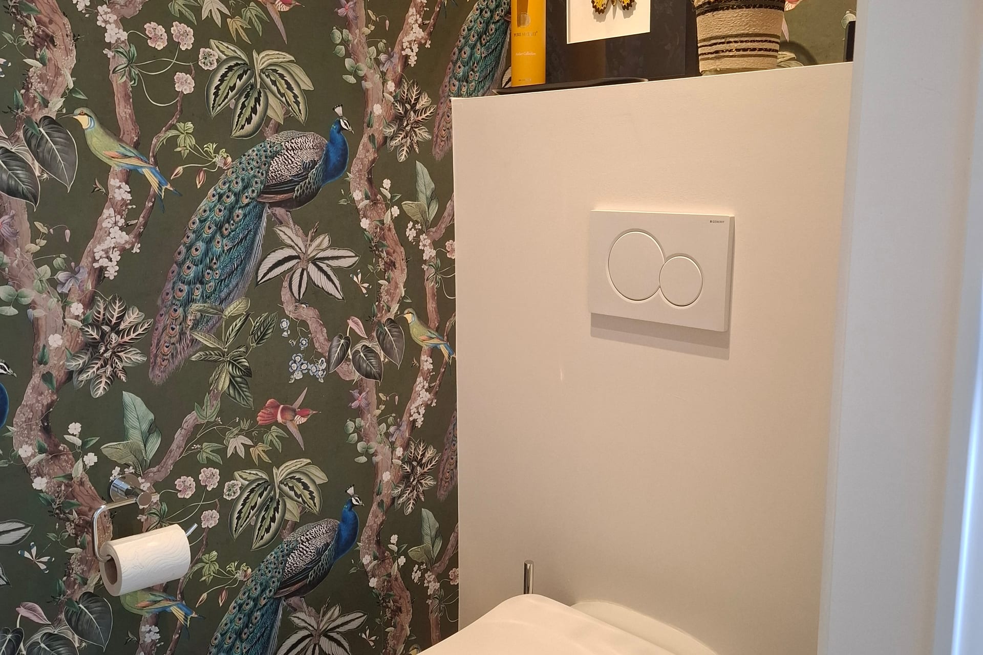 Plaque de commande Geberit à double touche sur un WC suspendu réparé par Atmos Technics à Bruxelles, mur tapissé d'un papier peint à motifs de paons.